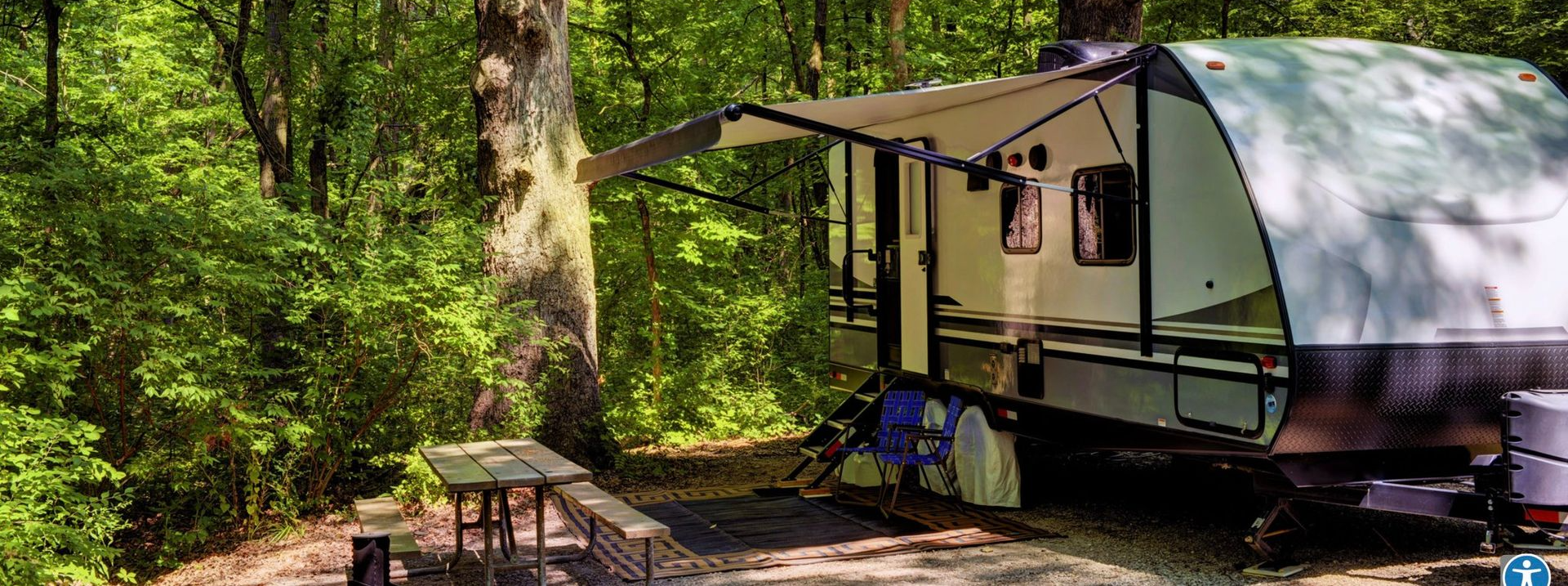 A camper trailer is parked in a wooded campsite with its awning extended and a picnic table nearby.