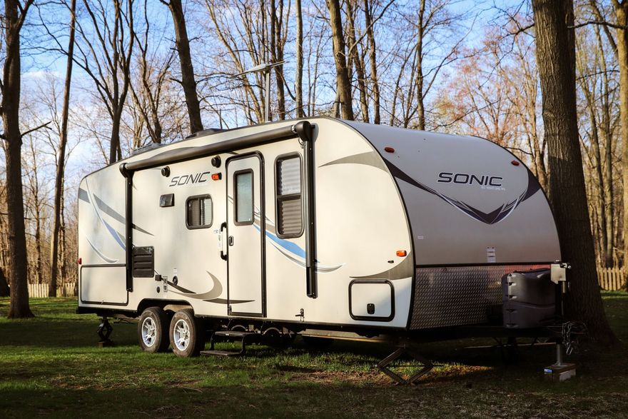 Travel trailer parked in a wooded area, featuring tan and gray exterior, awning extended.