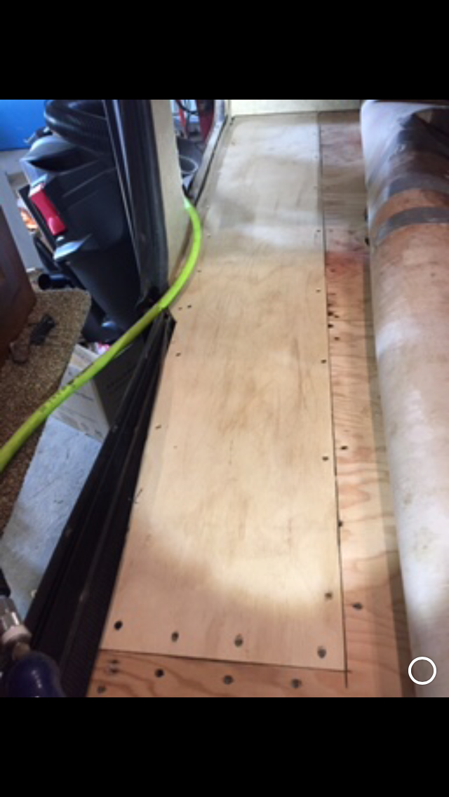 Plywood plank bolted to wooden structure, possibly a floor or platform inside a vehicle. A yellow hose is visible.