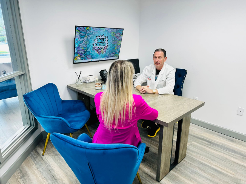 A person in pink sits across from a doctor in a white coat at a desk. The office has blue chairs.
