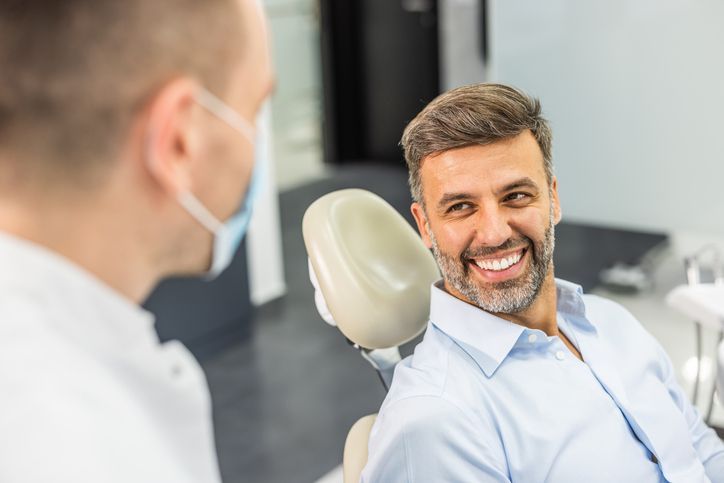 Man smiling at dentist in dental chair, blue shirt, neutral clinic background.