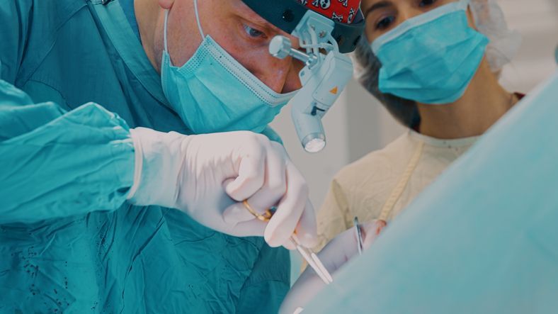 Surgeon in scrubs, mask, and headlamp, operating on a patient with an assistant in a surgical room.