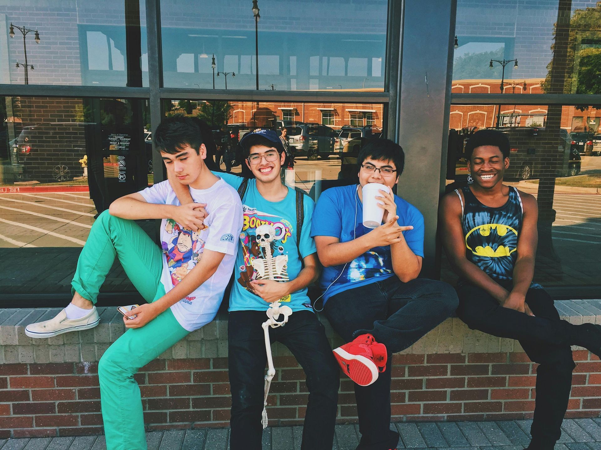 Four young men sitting on a ledge outside, one in teal pants, smiles, holding a drink, and a batman tank top.