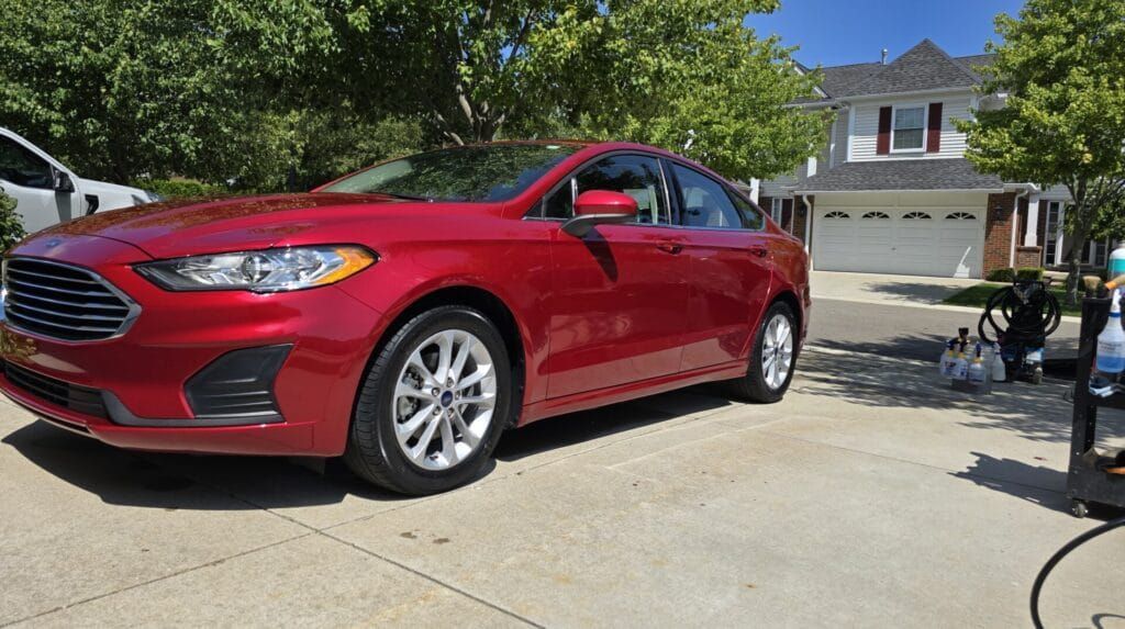 Red Ford sedan parked on a concrete driveway in front of a house on a sunny day.