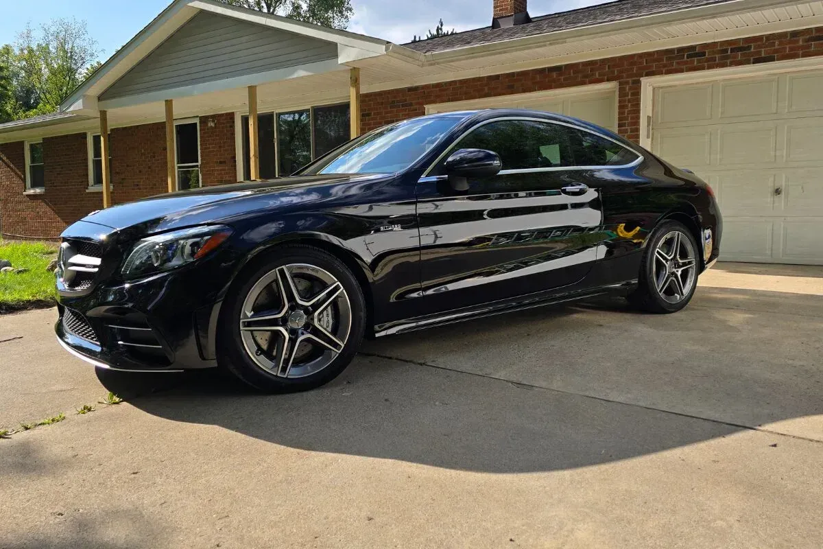 Black Mercedes coupe parked on a driveway in front of a brick house and garage on a sunny day.