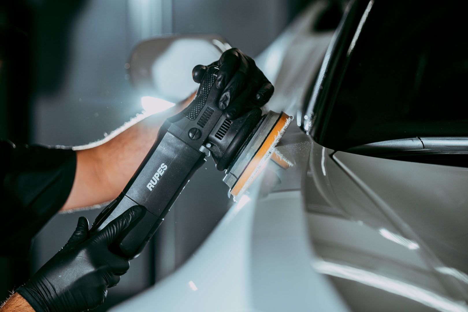 A person in black gloves polishing a white car with a black polisher in a garage setting.
