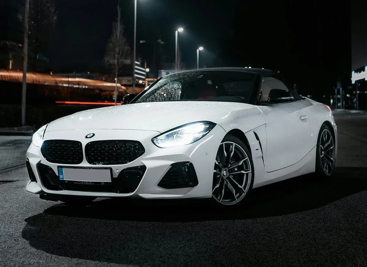 White BMW Z4 sports car parked on a dark city street at night, headlights on.