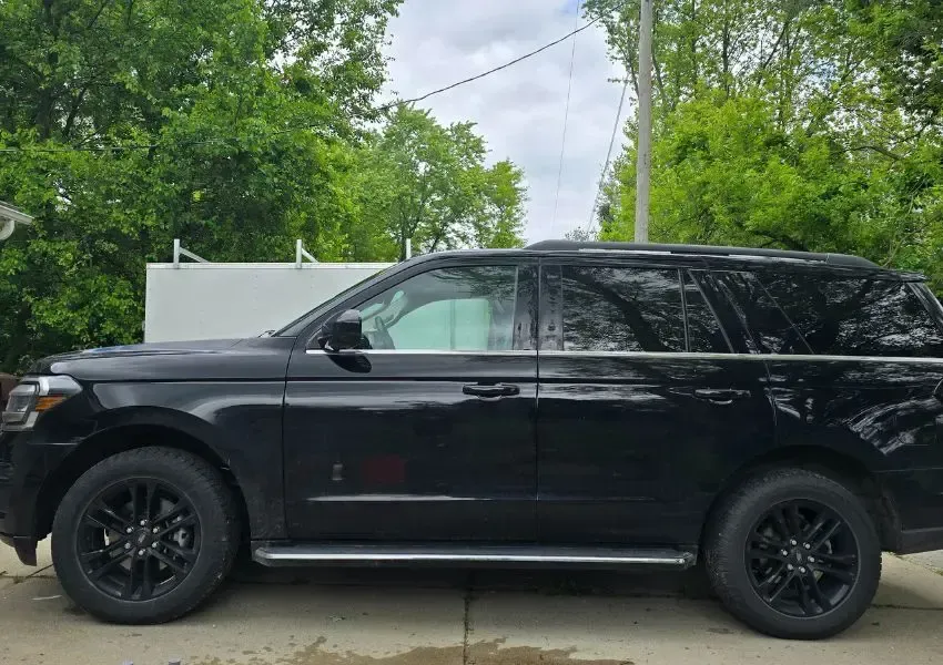 Black Ford Expedition parked outside, with black rims, against a backdrop of green trees and a cloudy sky.