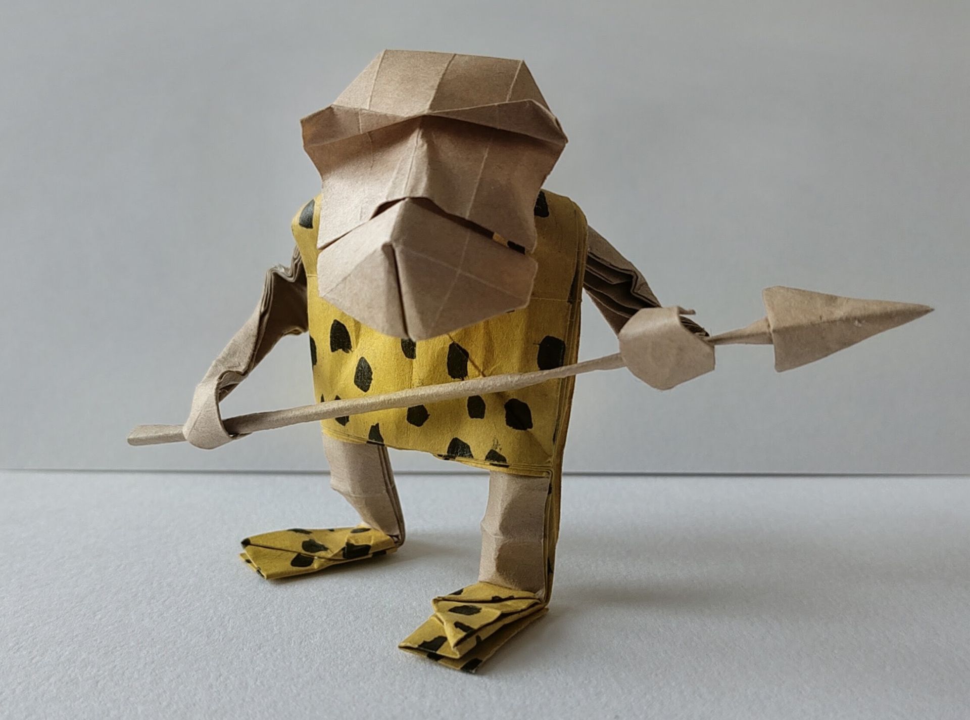 An origami apeman figure wearing a leopard skin vest and slippers and holding a spear
