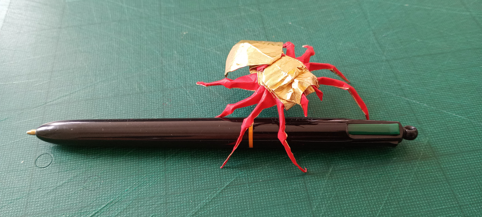 A golden origami spider with red legs seen walking over a pen