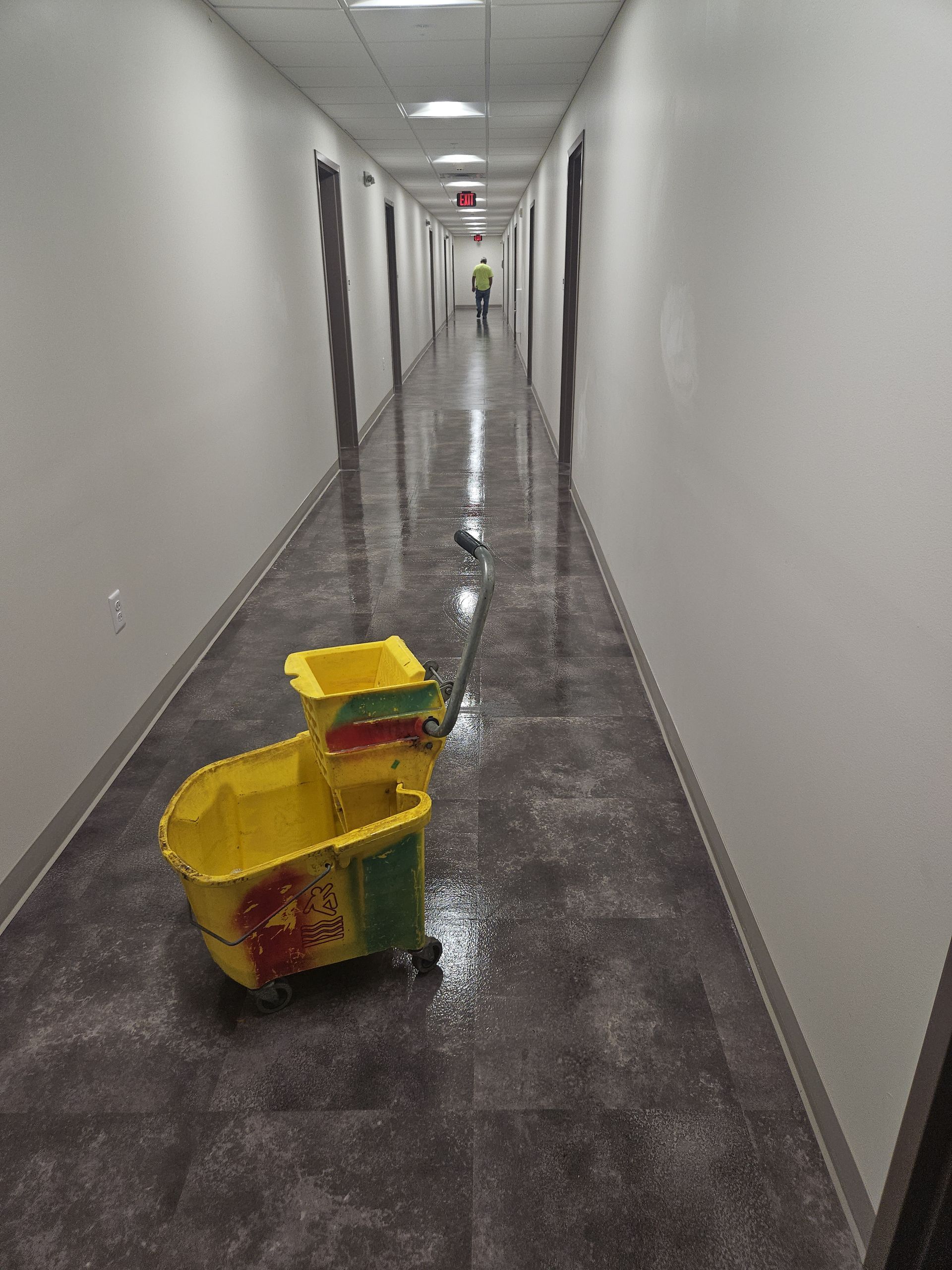 A person in teal scrubs mops a white floor in a bright, modern hospital hallway next to a yellow cleaning cart.