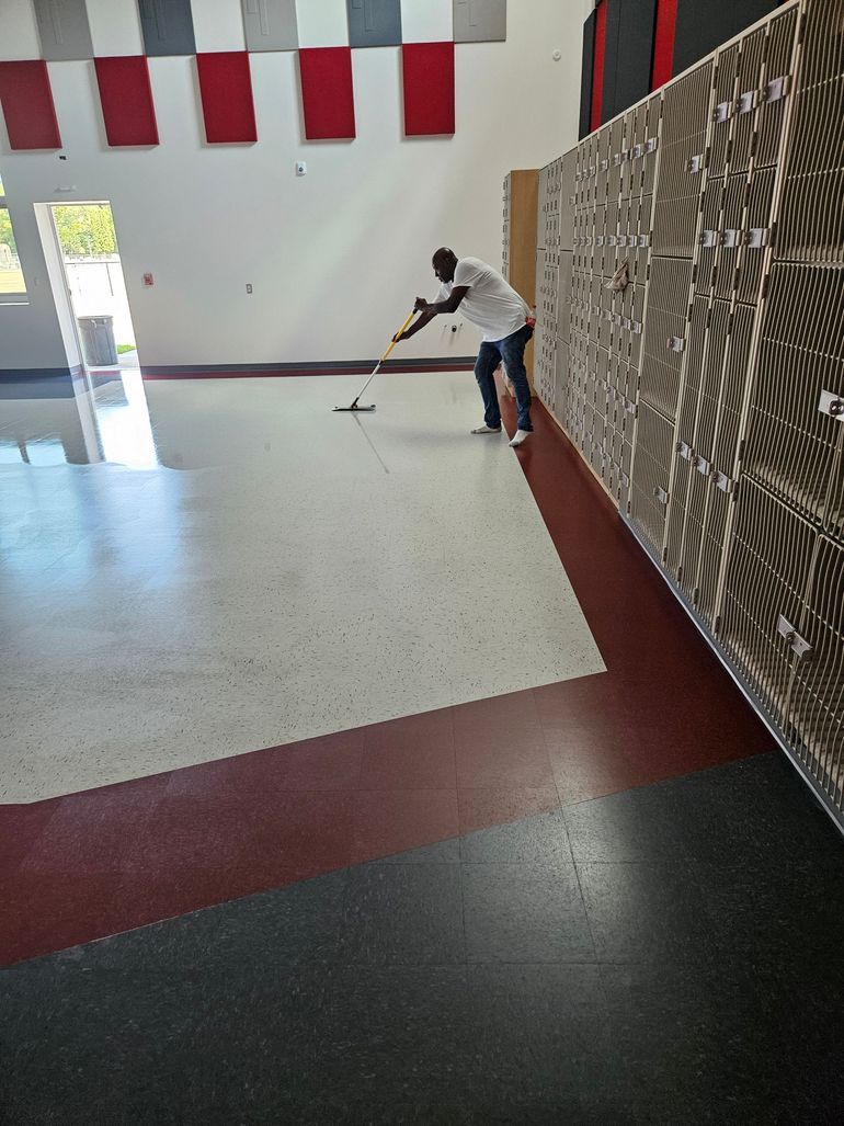 A person in a white shirt mops a speckled floor in a large room with lockers and rectangular wall accents.
