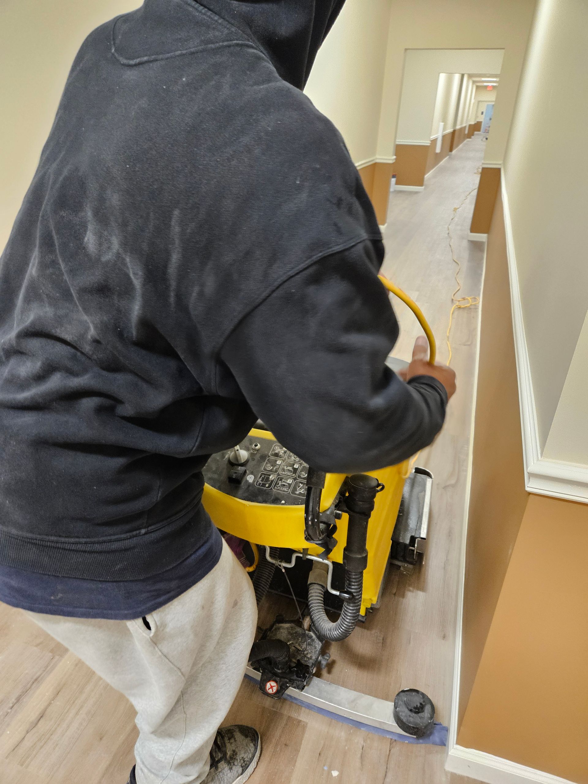 A yellow industrial floor scrubber sits on a polished marble floor in a spacious, modern hall with decorative columns.