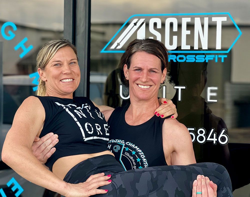 Two smiling women pose outside Ascent CrossFit in Hesperia, CA, one holding the other in her arms.