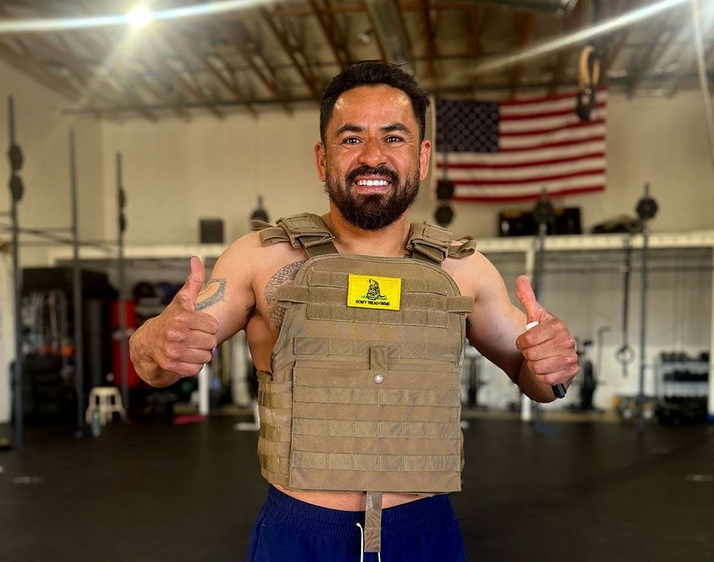 Man smiling at Ascent CrossFit in Hesperia, CA, wearing a tan tactical vest and giving two thumbs up, with an American flag behind him.