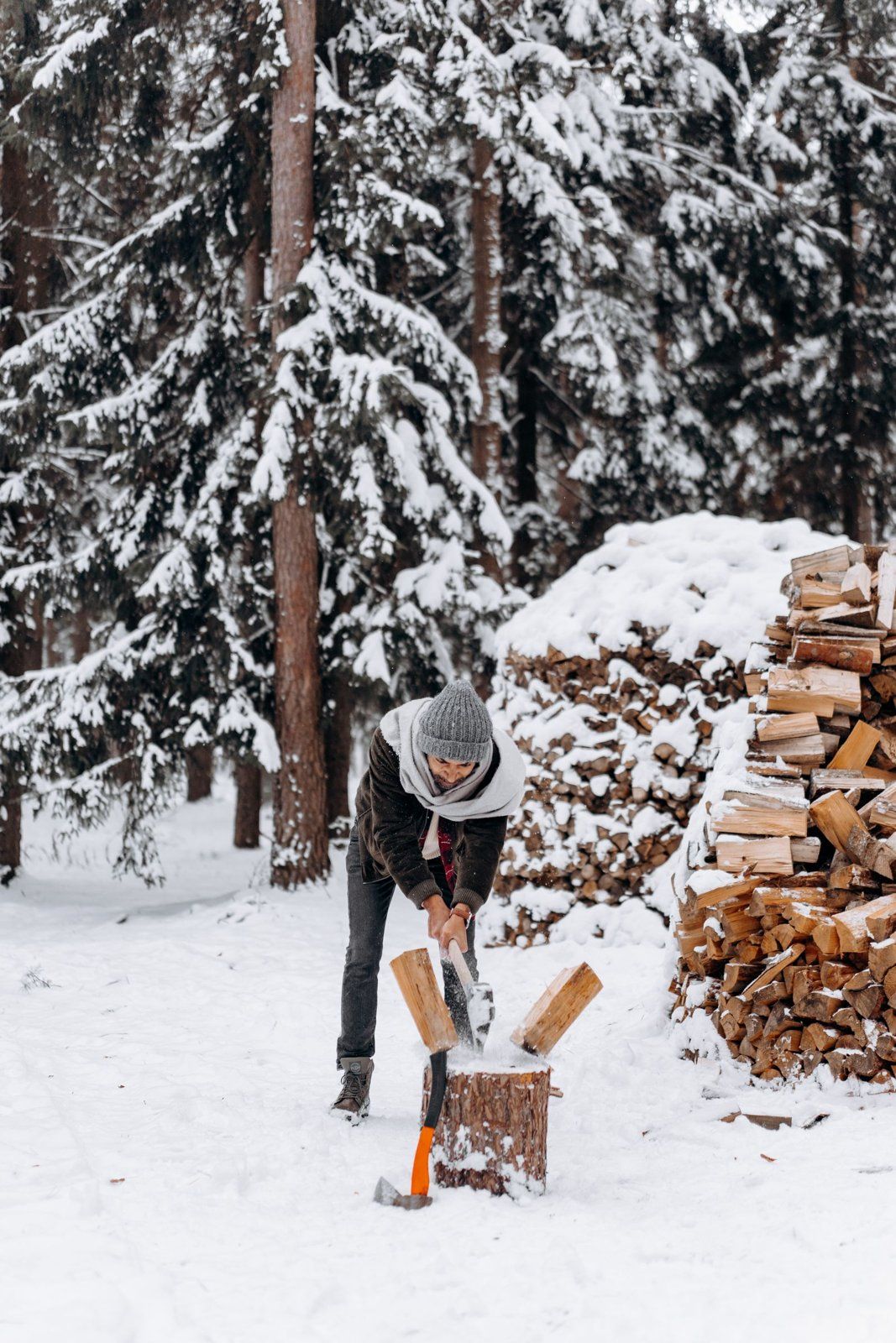 Person splitting firewood with an axe in a snowy yard next to stacked wood