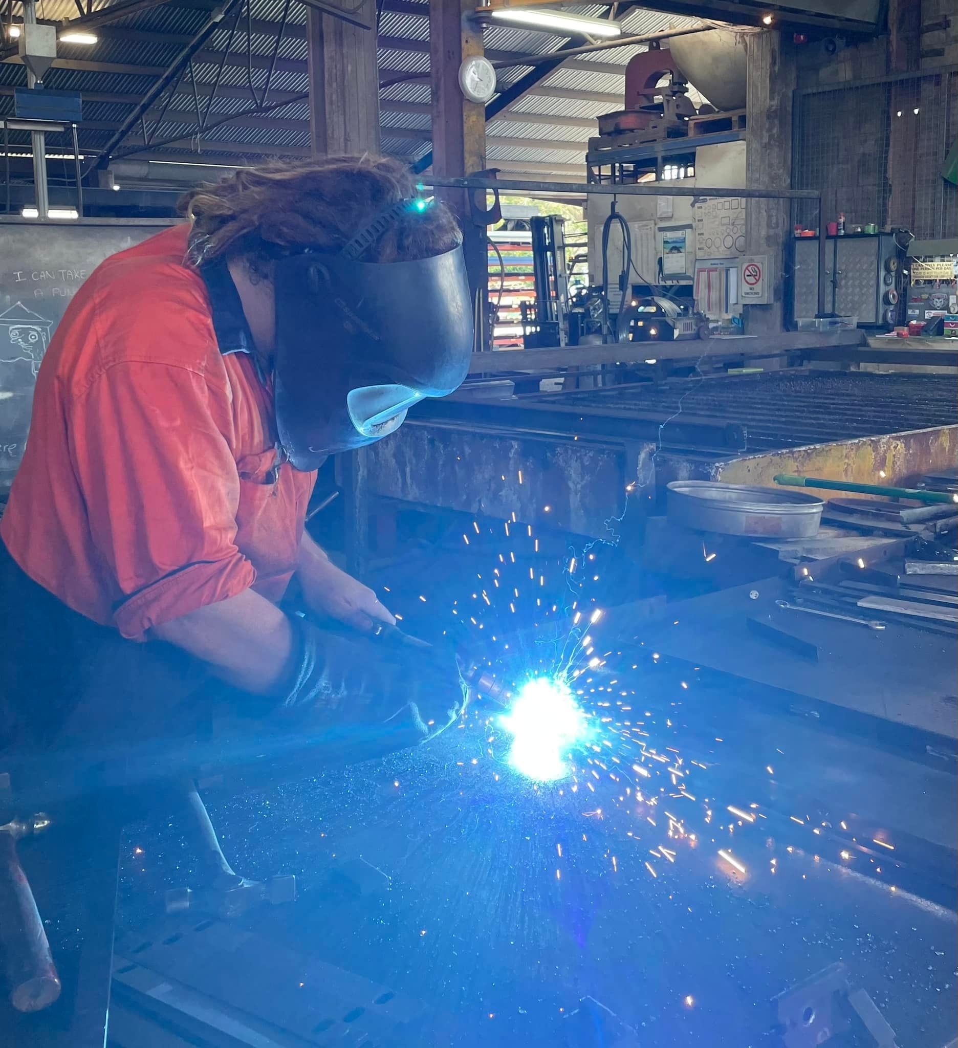 A Person in a Bright Orange Shirt and Welding Helmet Welds Metal — Ingham Agencies in Ingham, QLD