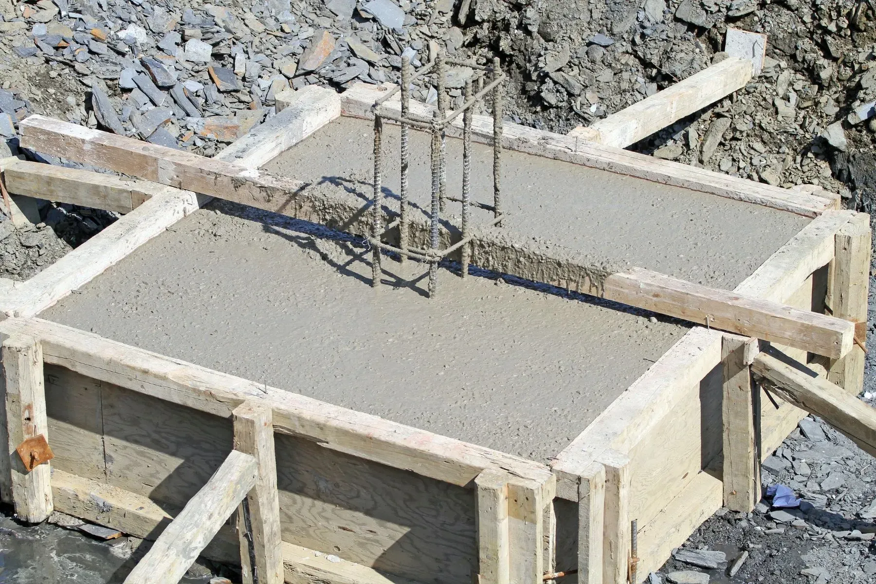 Freshly poured concrete footing with wooden formwork and exposed steel reinforcement bars at a construction site.