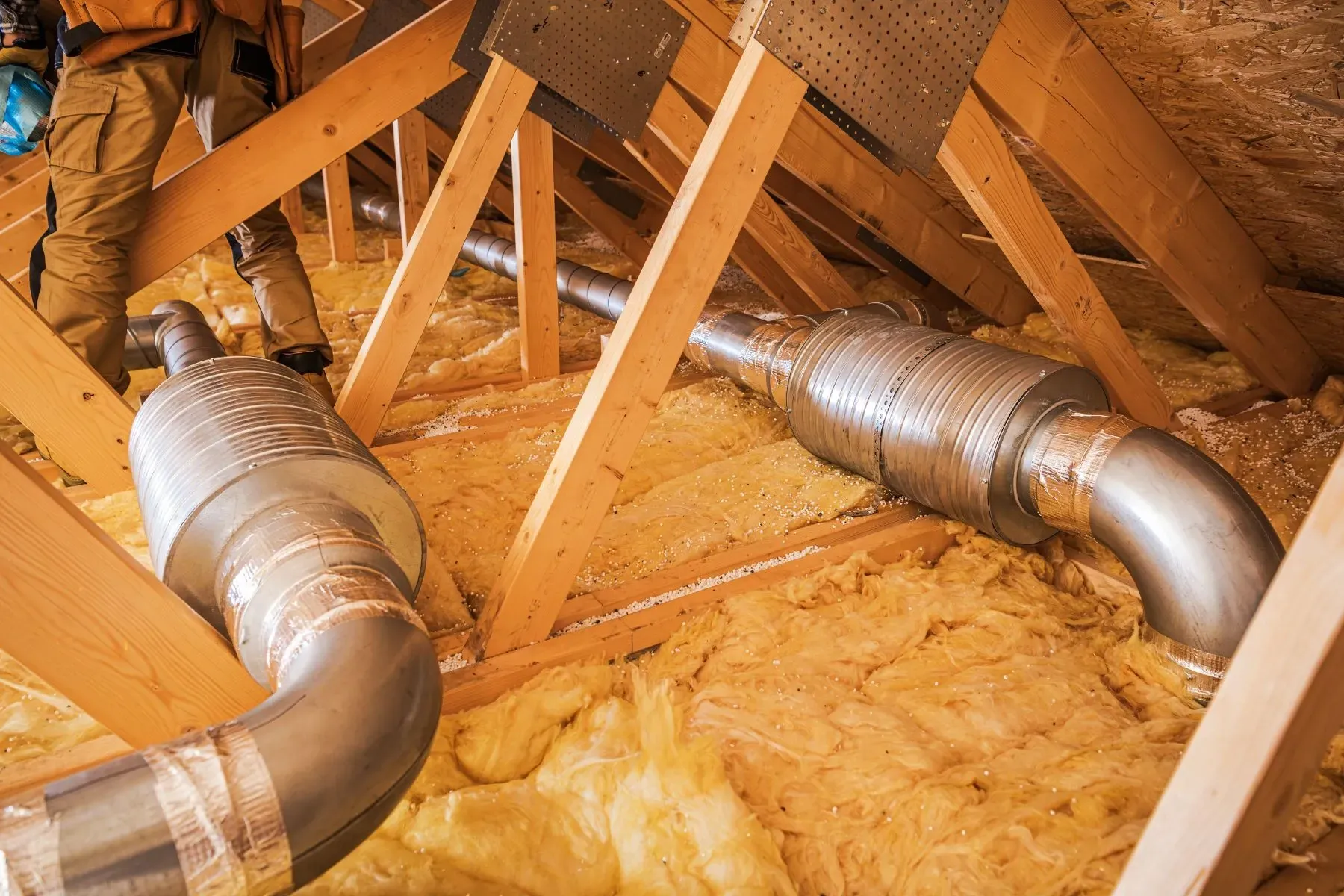 HVAC ductwork installation in an attic with fiberglass insulation and wooden roof trusses during home construction.