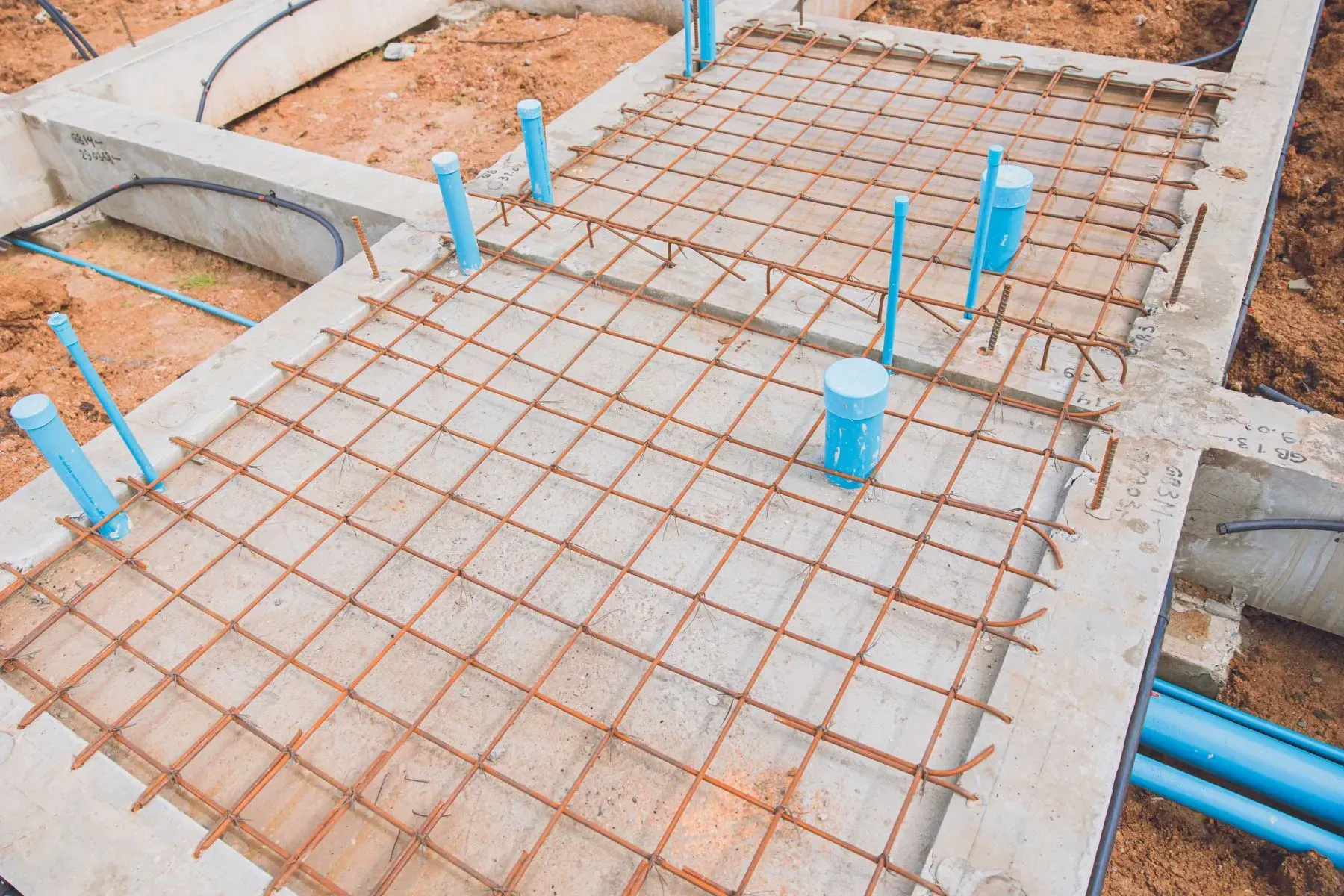 Concrete foundation with steel reinforcement grid and blue PVC plumbing pipes at a construction site showing early-stage building infrastructure work
