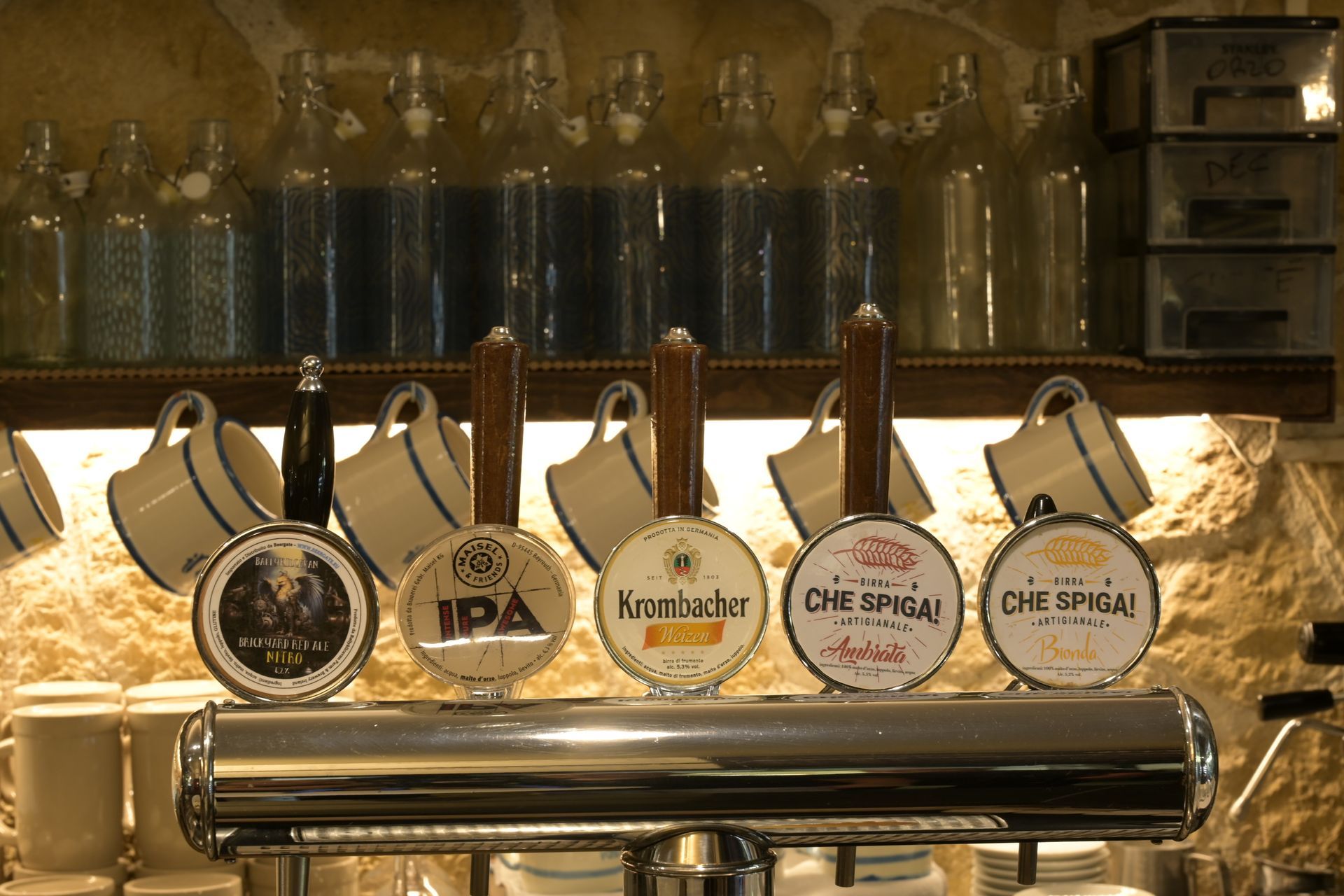 Spine di birra con etichette di birrifici assortiti, allineate su un bancone lucido, dietro uno scaffale illuminato con bottiglie