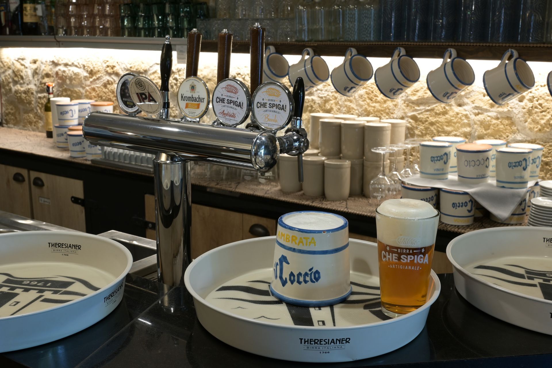 Postazione caffè con tazze, mug e piattini allineati sul bancone, sotto mug appesi e barattoli di condimenti