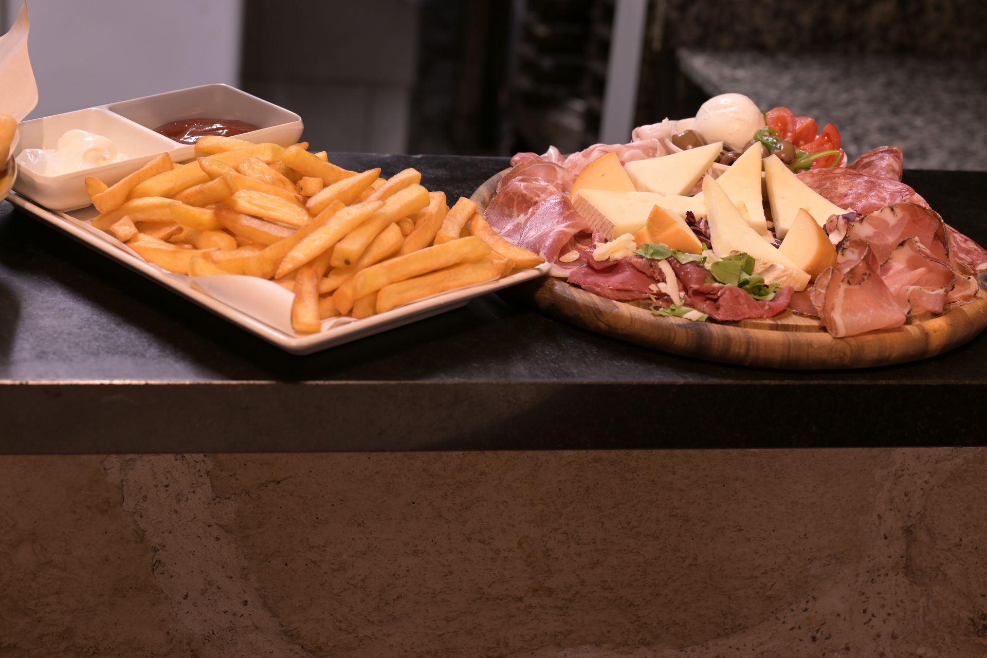 Due vassoi di cibo su un bancone: patatine fritte con salsa e un tagliere di formaggi e salumi