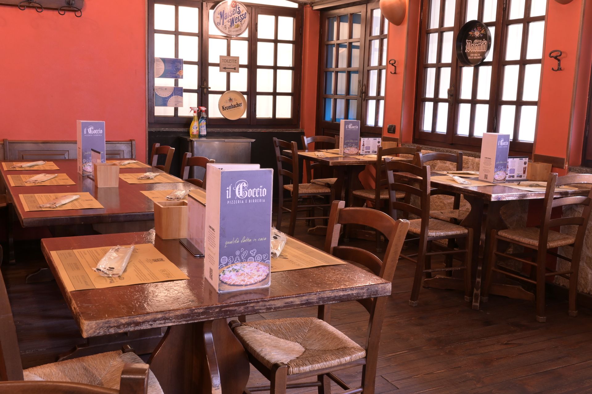 Sala da pranzo vuota di un ristorante, con tavoli e sedie in legno e portamenù vicino a grandi finestre.