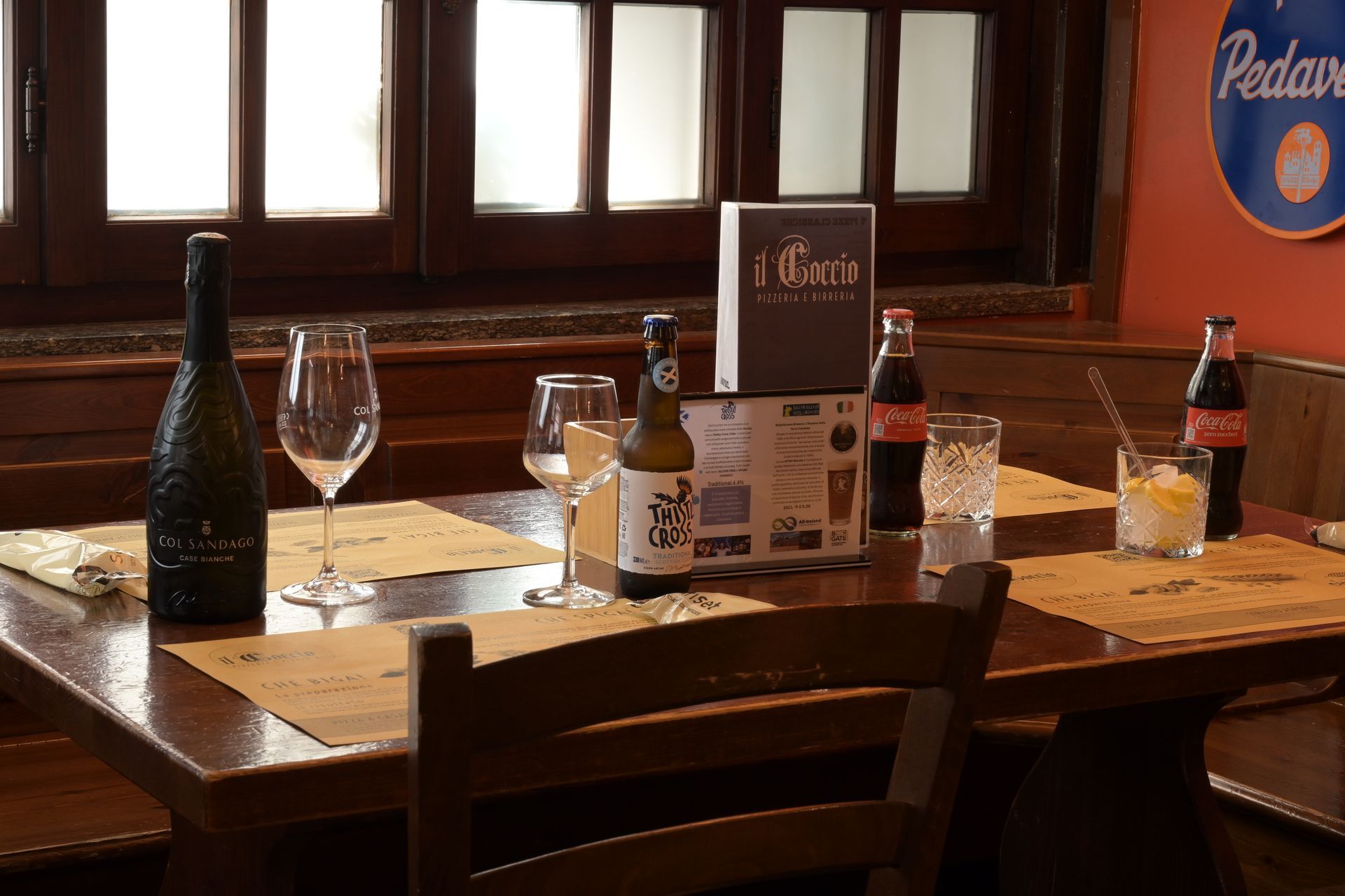 Tavolo di un ristorante poco illuminato, con bottiglie di birra, un calice di vino, menù e condimenti vicino a una finestra