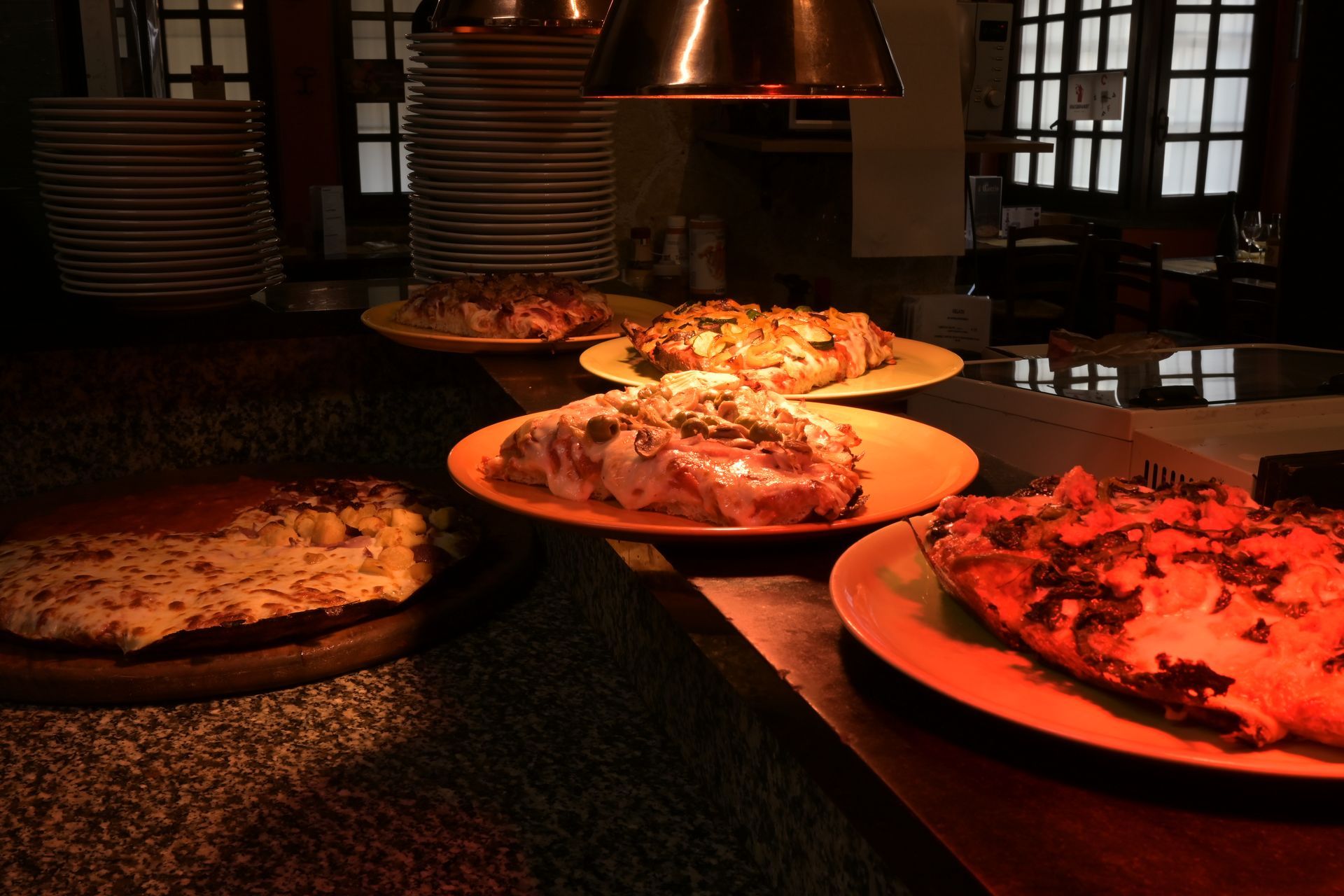 Diverse pizze su vassoi riscaldati sotto lampade riscaldanti in un buffet di un ristorante poco illuminato