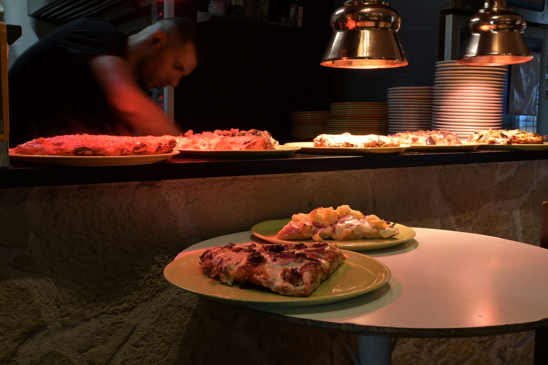 Piatti di cibo su un bancone da buffet sotto lampade riscaldanti dalla luce calda, con una persona sfocata che si serve sullo sfondo