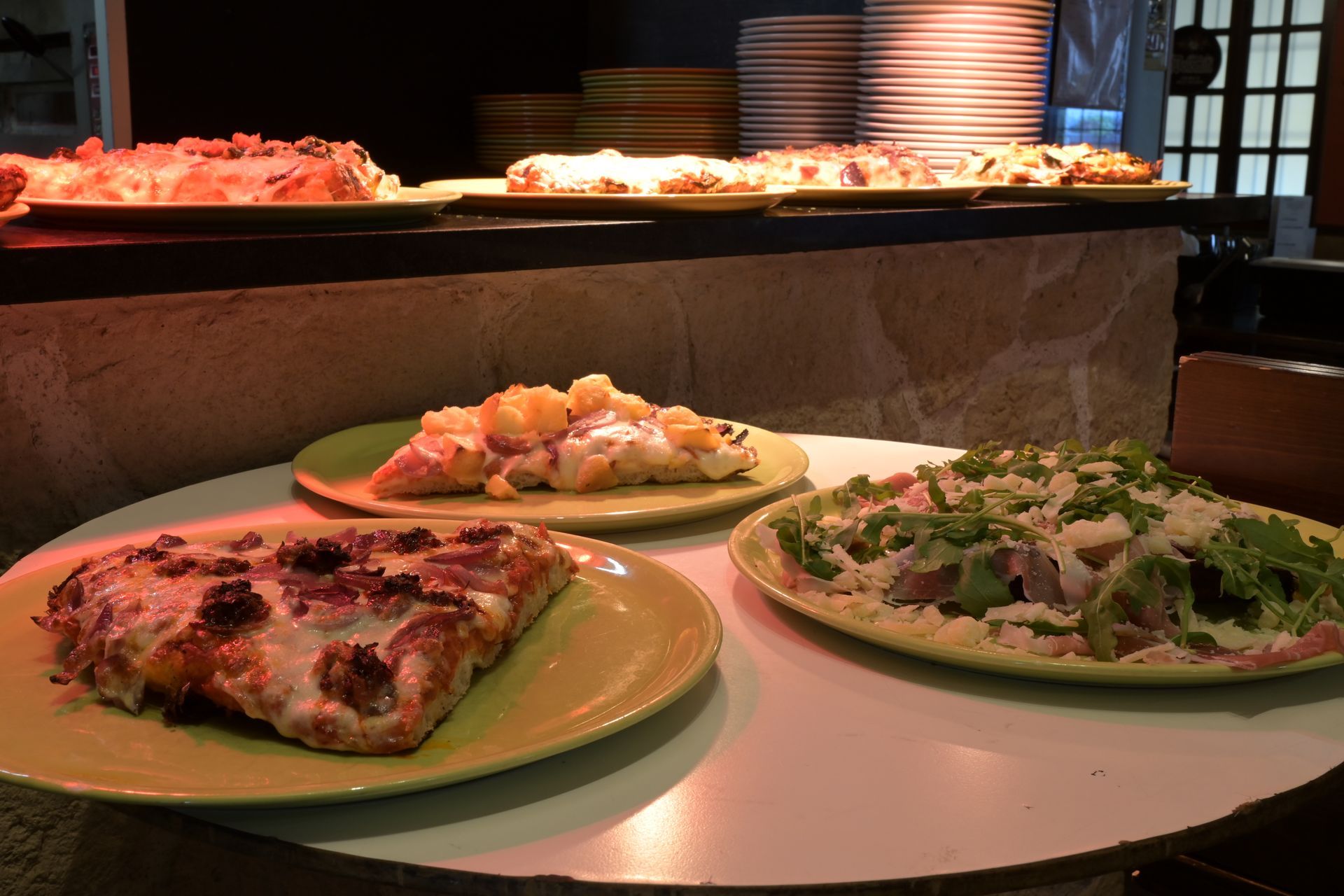 Tre pizze servite su un tavolo al banco di un buffet, con un’illuminazione calda