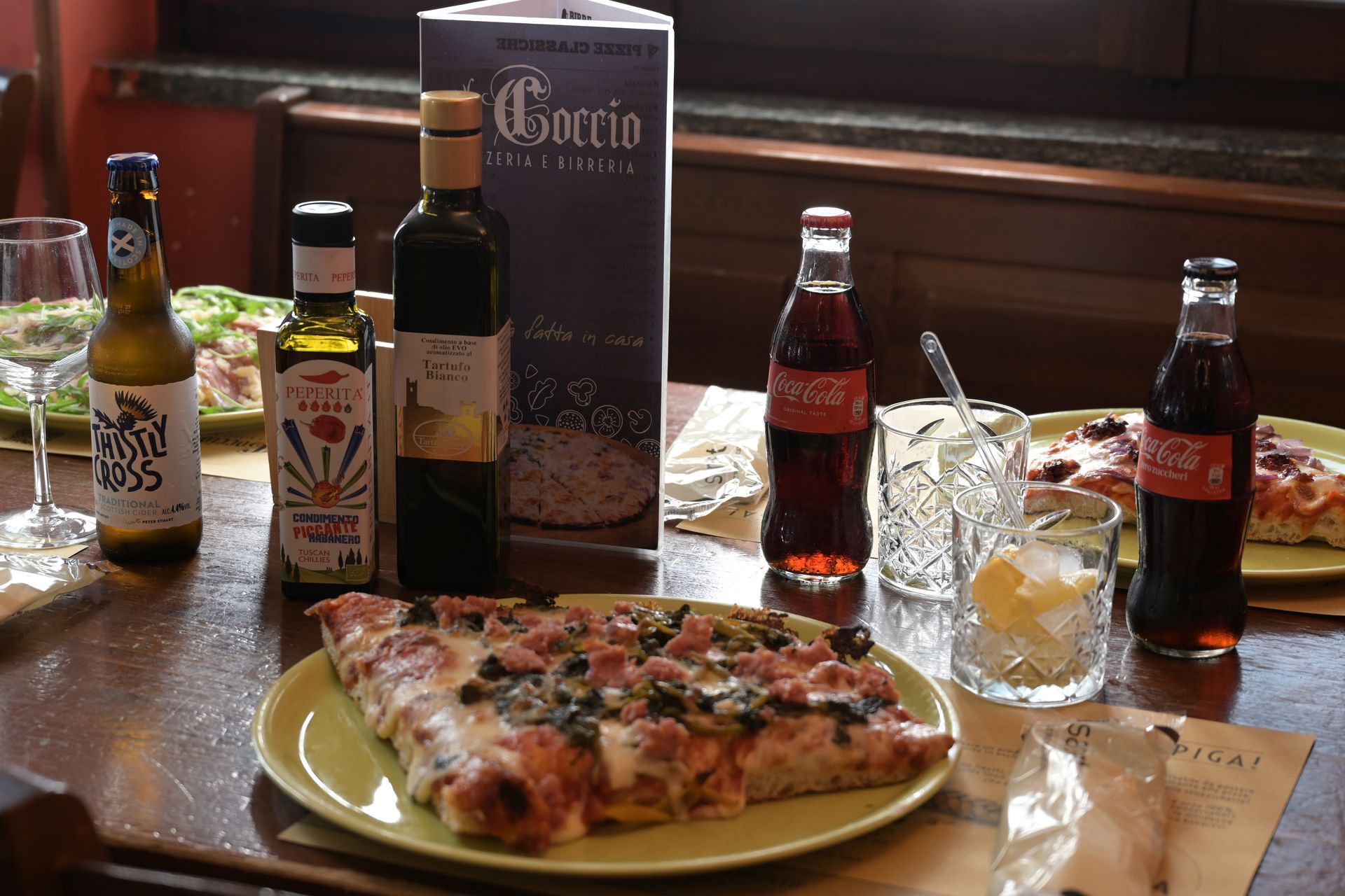 Tavolo di un ristorante con pizza, bevande, condimenti e un menu piegato, in penombra