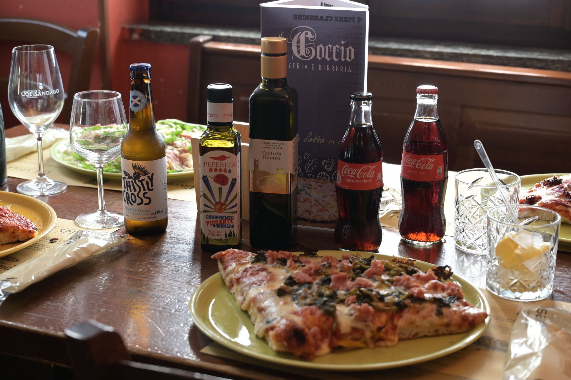 Tavolo apparecchiato con pizza, bevande, bottiglie di vino e un menù in una saletta di ristorante