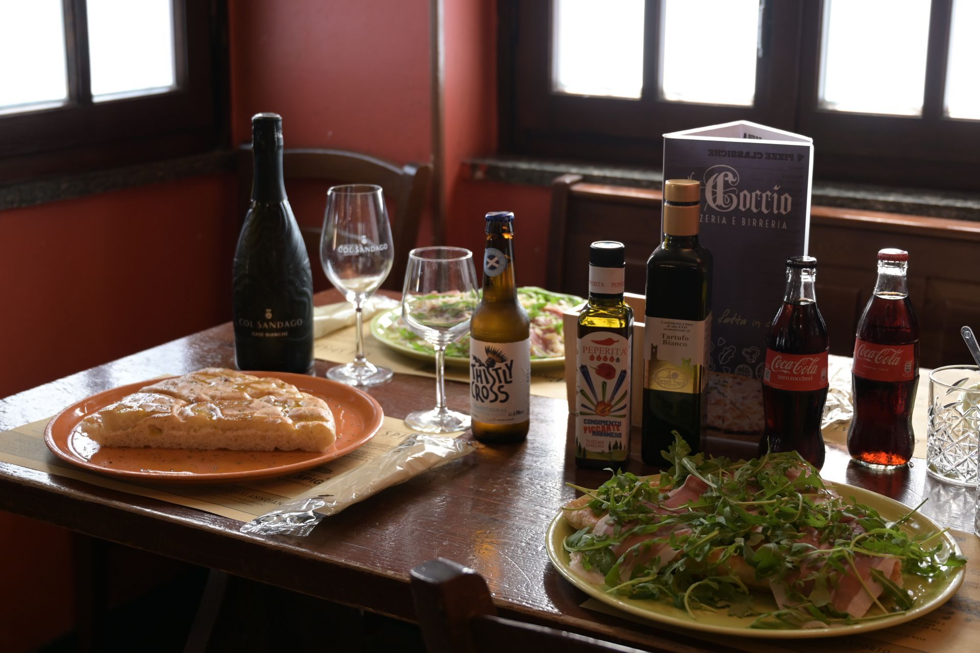 Un tavolo con pane, insalata, bevande e bottiglie di condimenti vicino a una finestra in un ristorante.