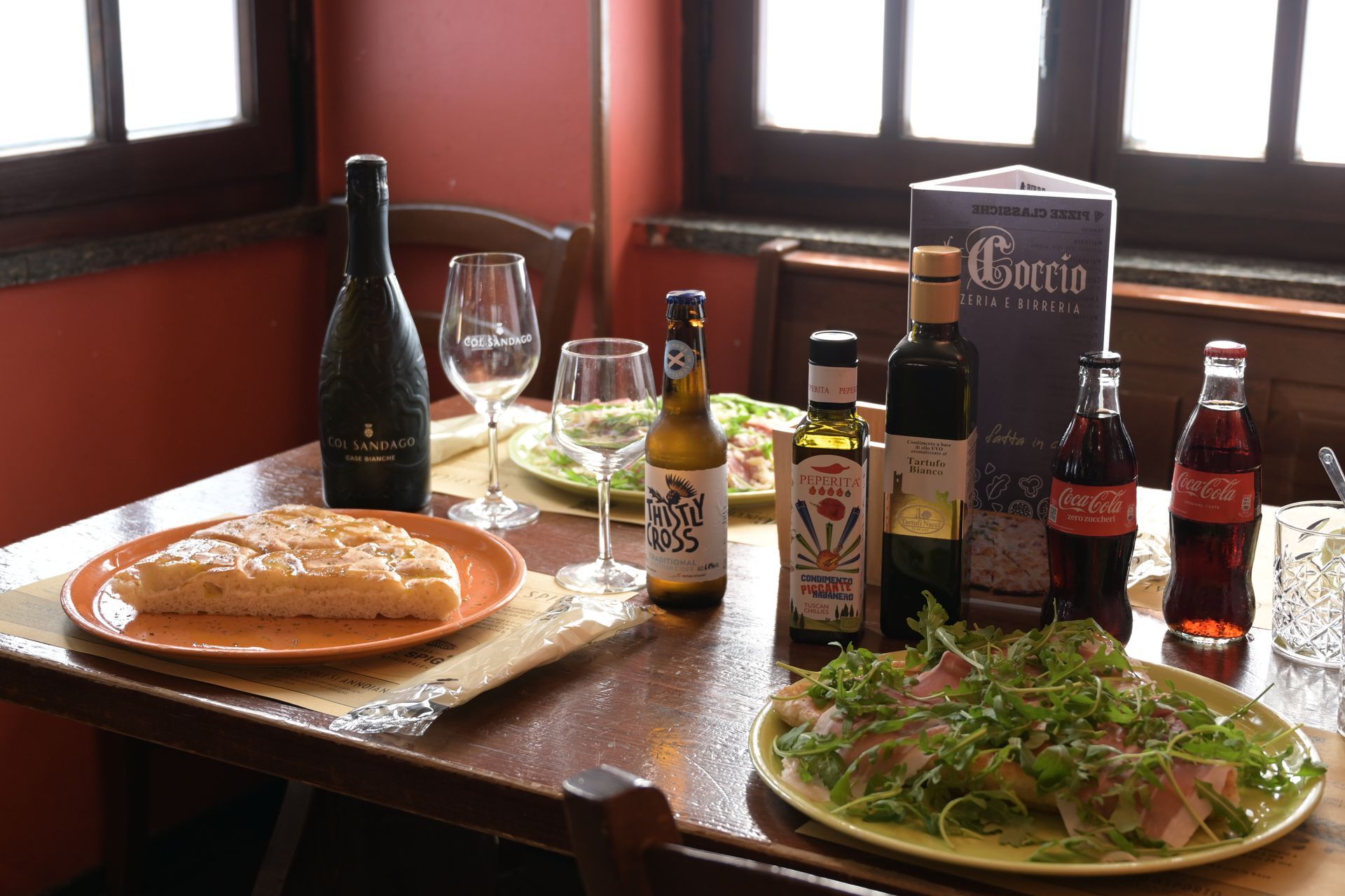 Tavolo da pranzo con pizza, insalata, calici di vino e bottiglie vicino a una finestra.