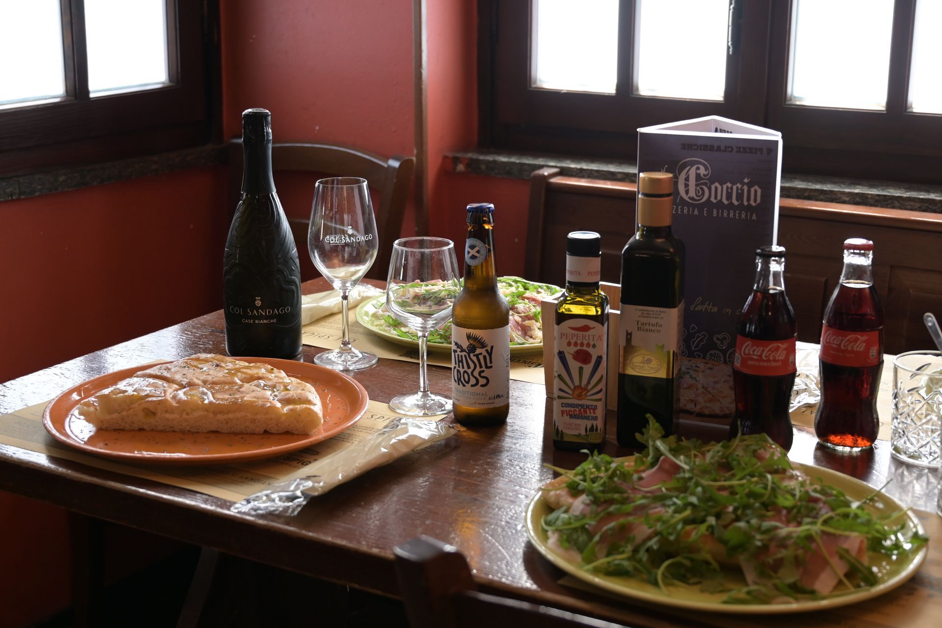 Tavolo di un ristorante con pizza, insalata, vino e una varietà di bevande in bottiglia vicino a una finestra