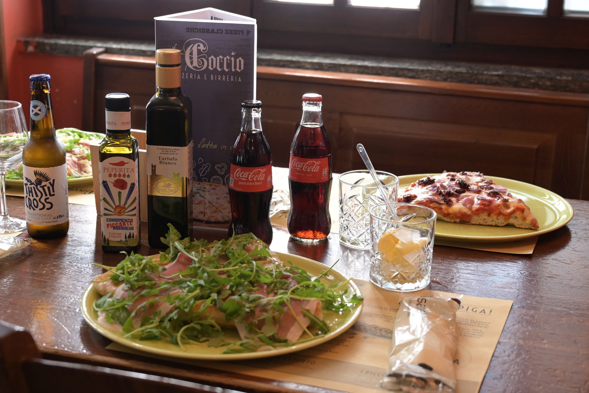 Tavolo di un ristorante con insalata, pizza, bevande e bottiglie di vino vicino a una finestra