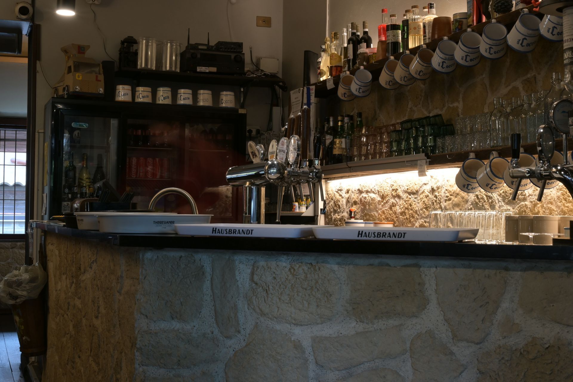 Bancone del bar con spillatori di birra, calici da vino appesi e bottiglie in un ambiente accogliente e illuminato da luci calde