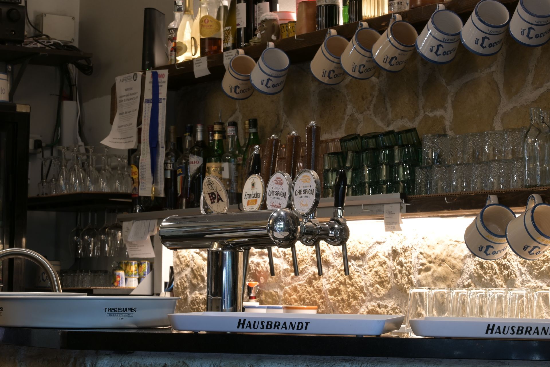 Bar con file di tazze appese, bottiglie, bicchieri e una spillatrice per birra sotto luci calde