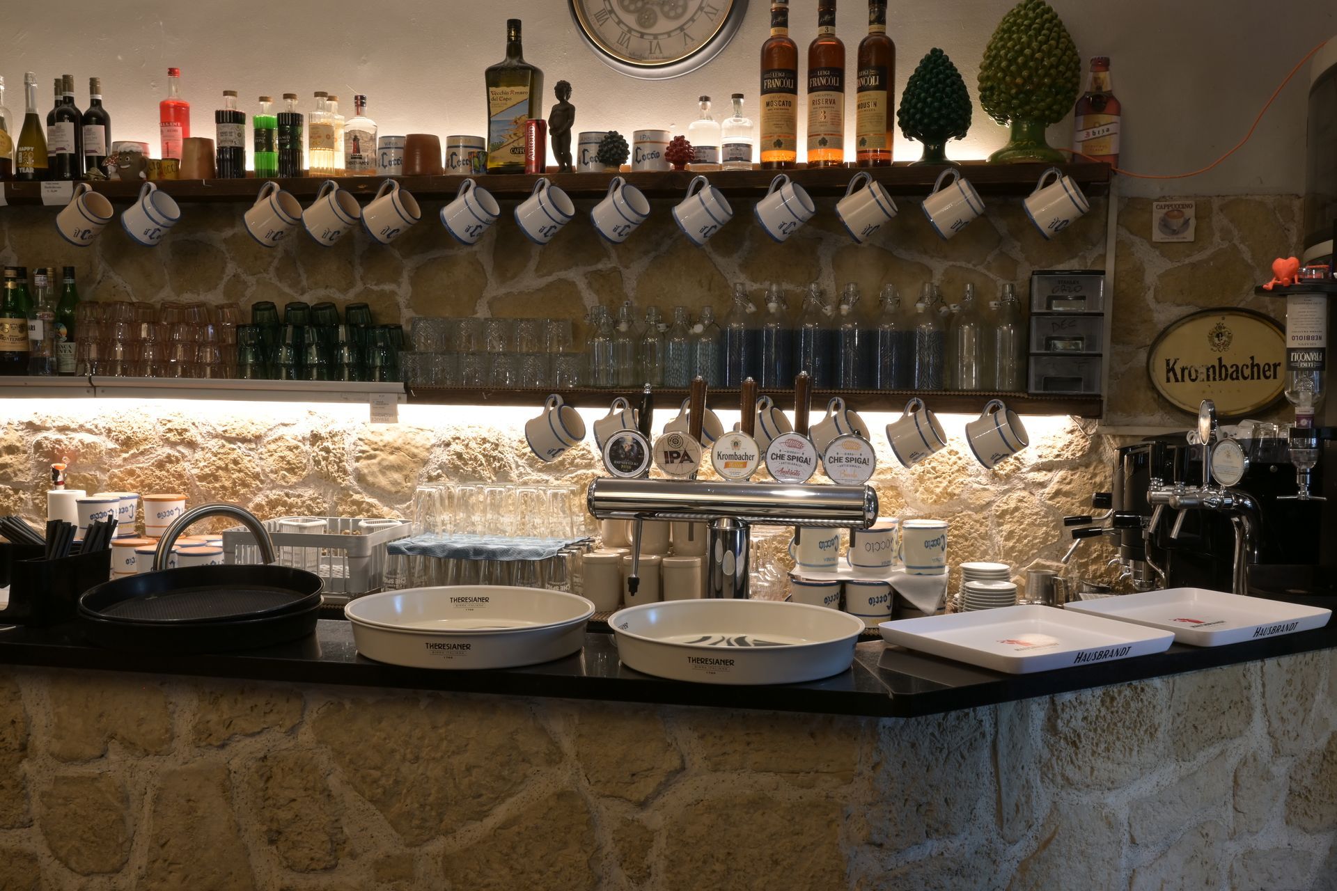 Bancone di enoteca con bicchieri appesi, bottiglie e tazze sotto una luce calda