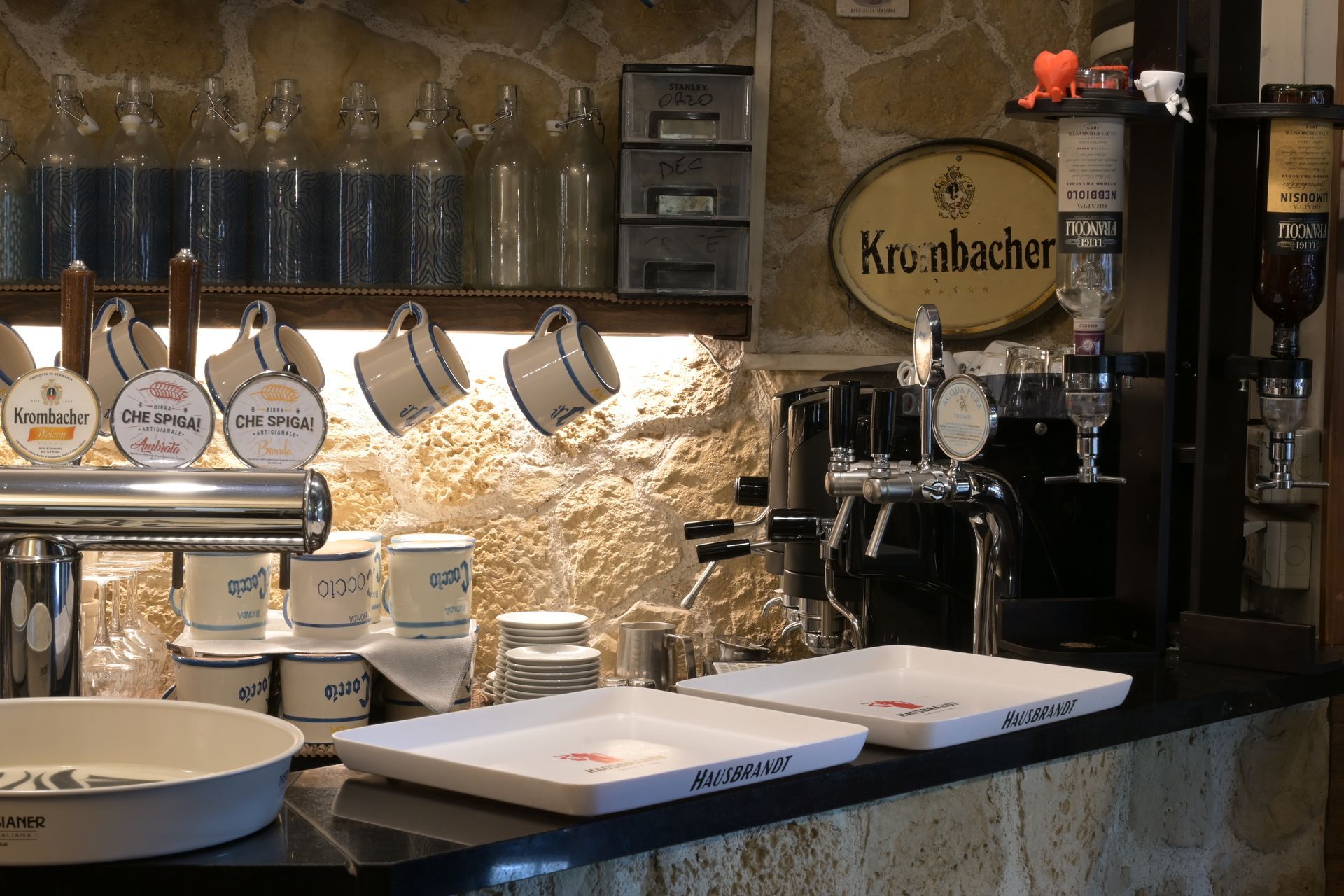 Bar con spillatori di birra, bottiglie e bicchieri su un bancone di pietra, in un ambiente rustico