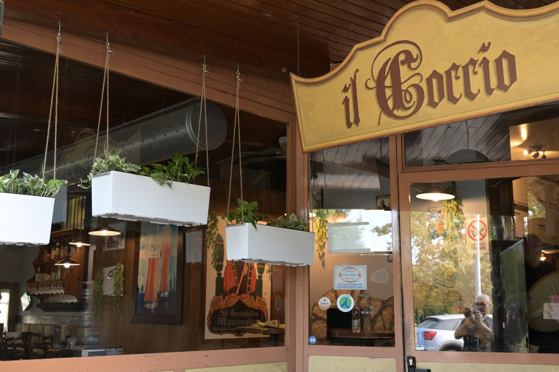 Ingresso del ristorante con un’insegna di legno con la scritta “il Coccio” e fioriere bianche sospese.