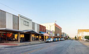 Sunlit street of colorful storefronts and warehouses with parked cars on an empty road