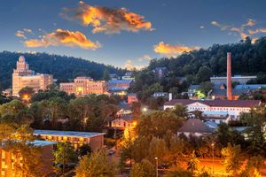 Dusk cityscape with glowing buildings, a tall chimney, and colorful clouds over wooded hills