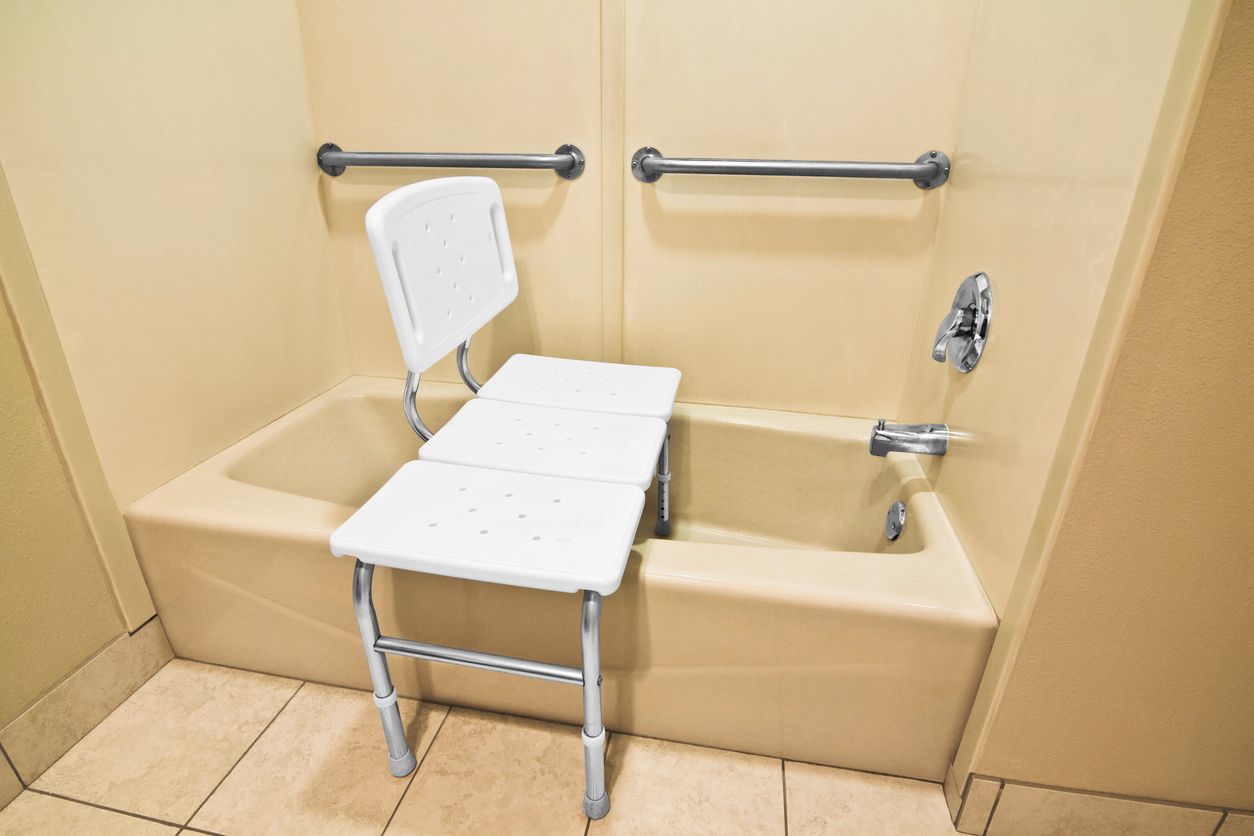 Accessible bathtub with a white shower chair, grab bars, and handheld shower in a beige bathroom