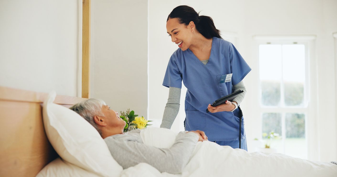Nurse in blue scrubs speaking with a patient in bed in a bright hospital room