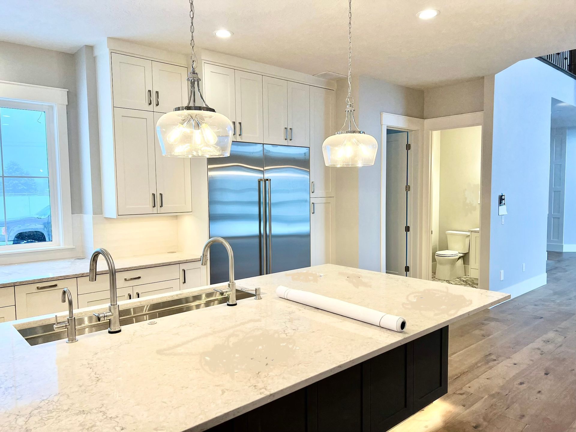 Modern kitchen with marble island, pendant lights, stainless fridge, and hardwood floors.