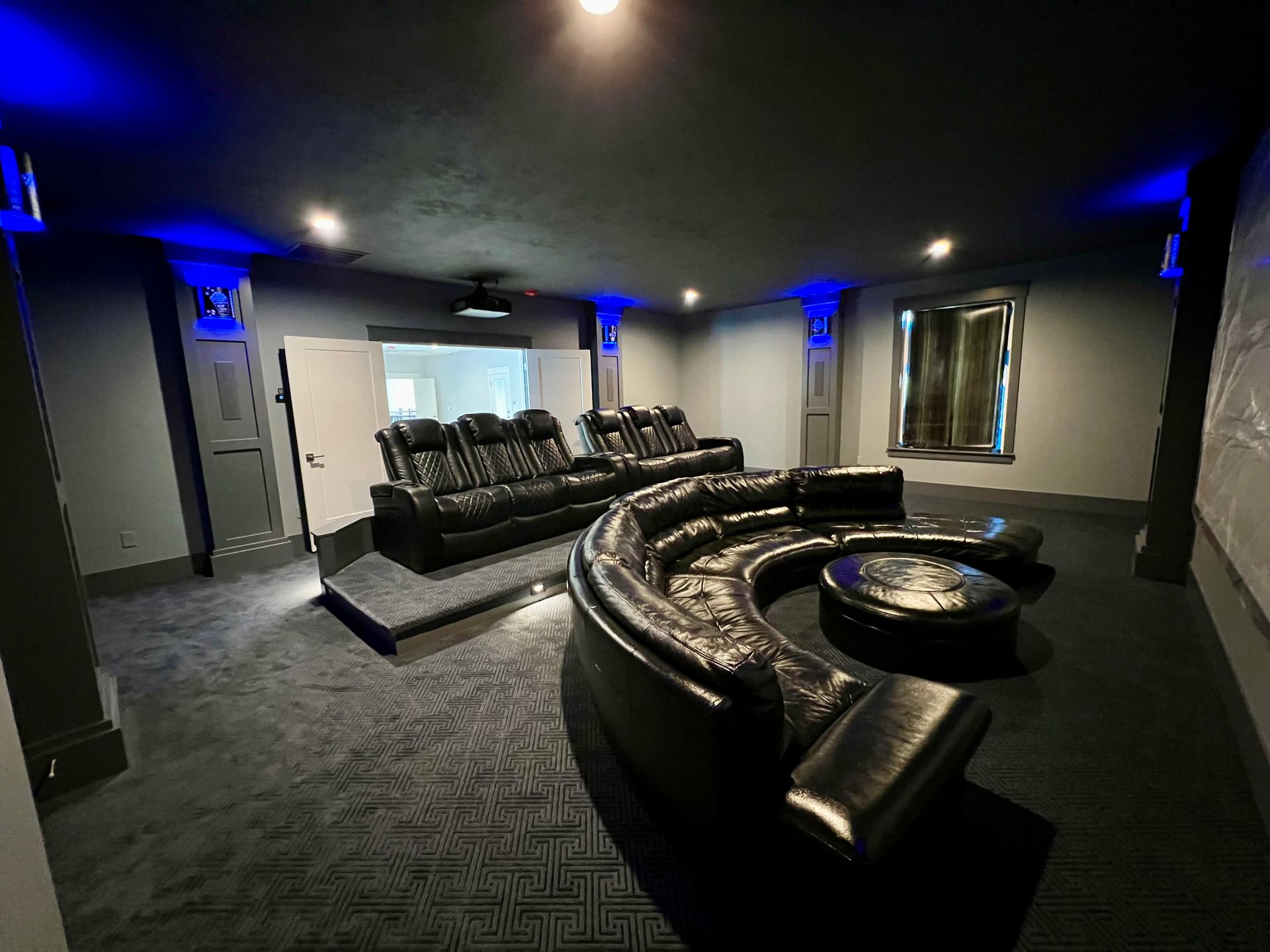 Modern home theater with black leather sectional seating, dark carpet, blue accent lighting, and wall sconces
