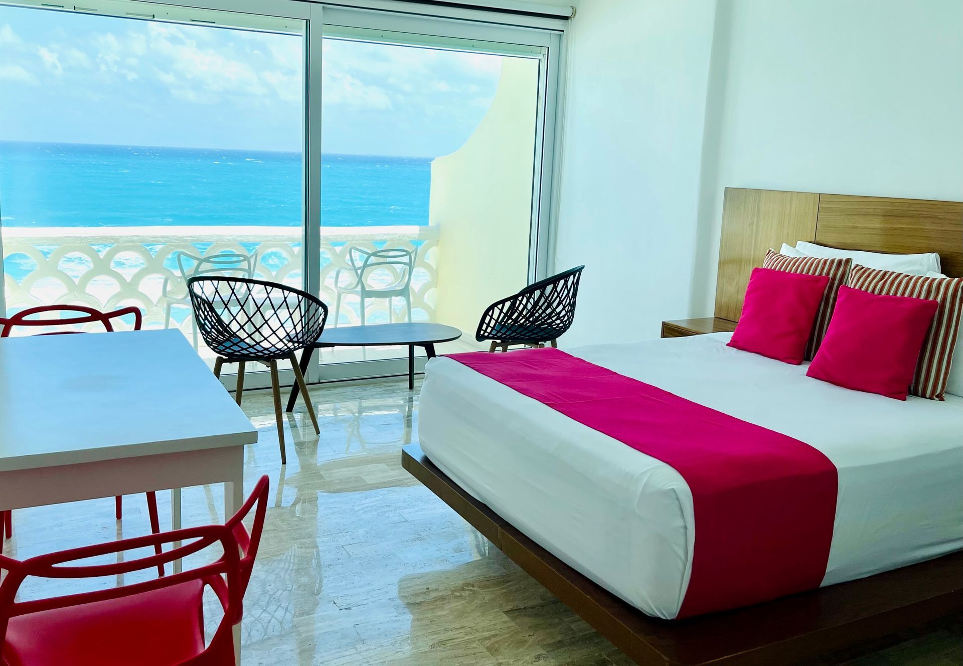 Dormitorio con vista al mar; cama con detalles en rosa, balcón con sillas, mesa blanca con sillas rojas.